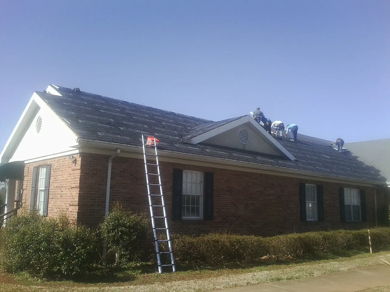 Crew tearing shingles off a brick ranch roof with a ladder set — Emergency Roof Repair work by our crew in Tryon